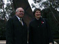 Two people in black coats standing together in front of handle shaped bronze memorial.